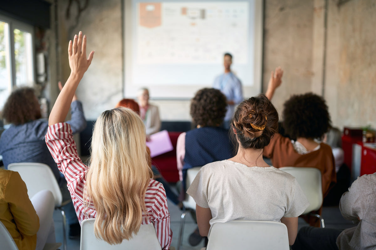 A classroom or seminar scene with several people seated and raising their hands toward a presenter standing in front of a projection screen.