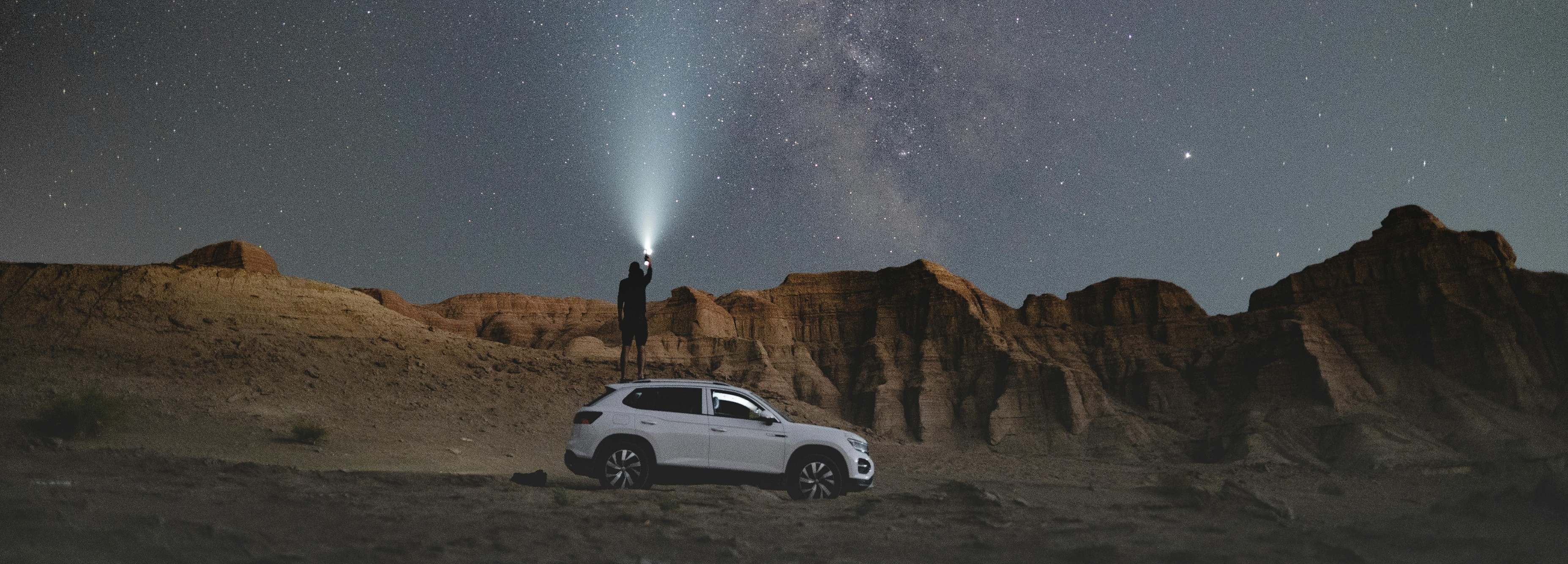 A man standing on a rocky hill in a desert, shining a bright flashlight beam up into the night sky filled with the Milky Way and stars, with a white SUV parked below him.