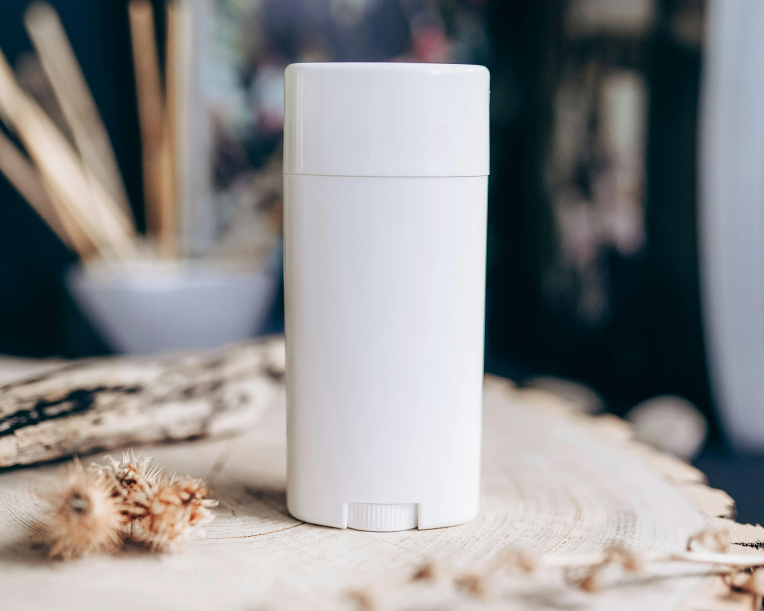 A shot of a white, unbranded deodorant stick sitting upright on a piece of light-colored, rustic-looking wood. In the background, there's a book and other natural, dried elements.