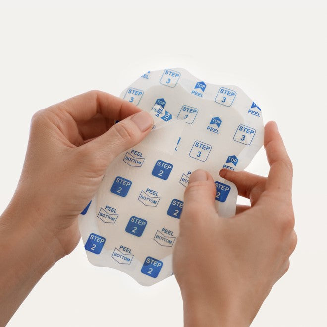 A close-up of hands peeling back Clear Inner Liner printed with blue and white 'STEP 2' and 'PEEL BOTTOM' labels, showing a translucent adhesive patch underneath on a clean white background.