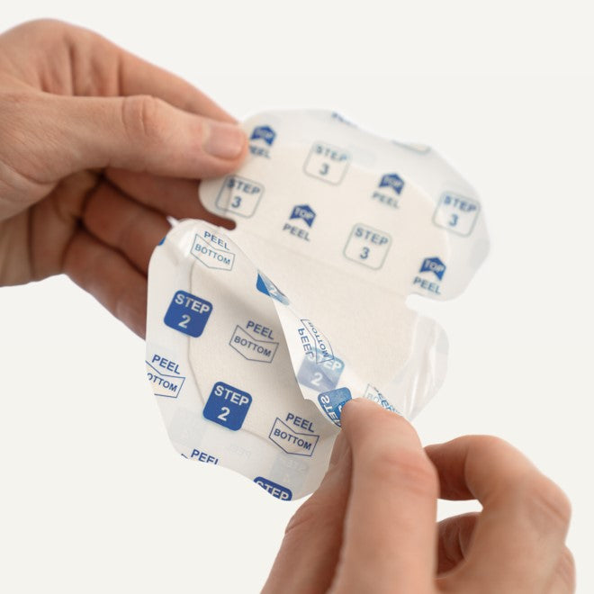 A close-up of hands peeling back Clear Inner Liner printed with blue and white 'STEP 2' and 'PEEL BOTTOM' labels, showing a translucent adhesive patch underneath on a clean white background.