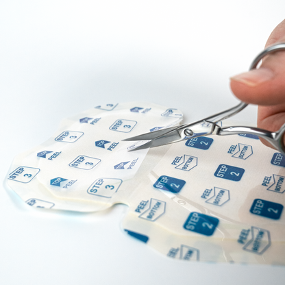 A close-up of a hand using metal scissors to cut the top half of a Clear Inner Liner of adhesive sweat protection patch labeled with step numbers on a clean white background.